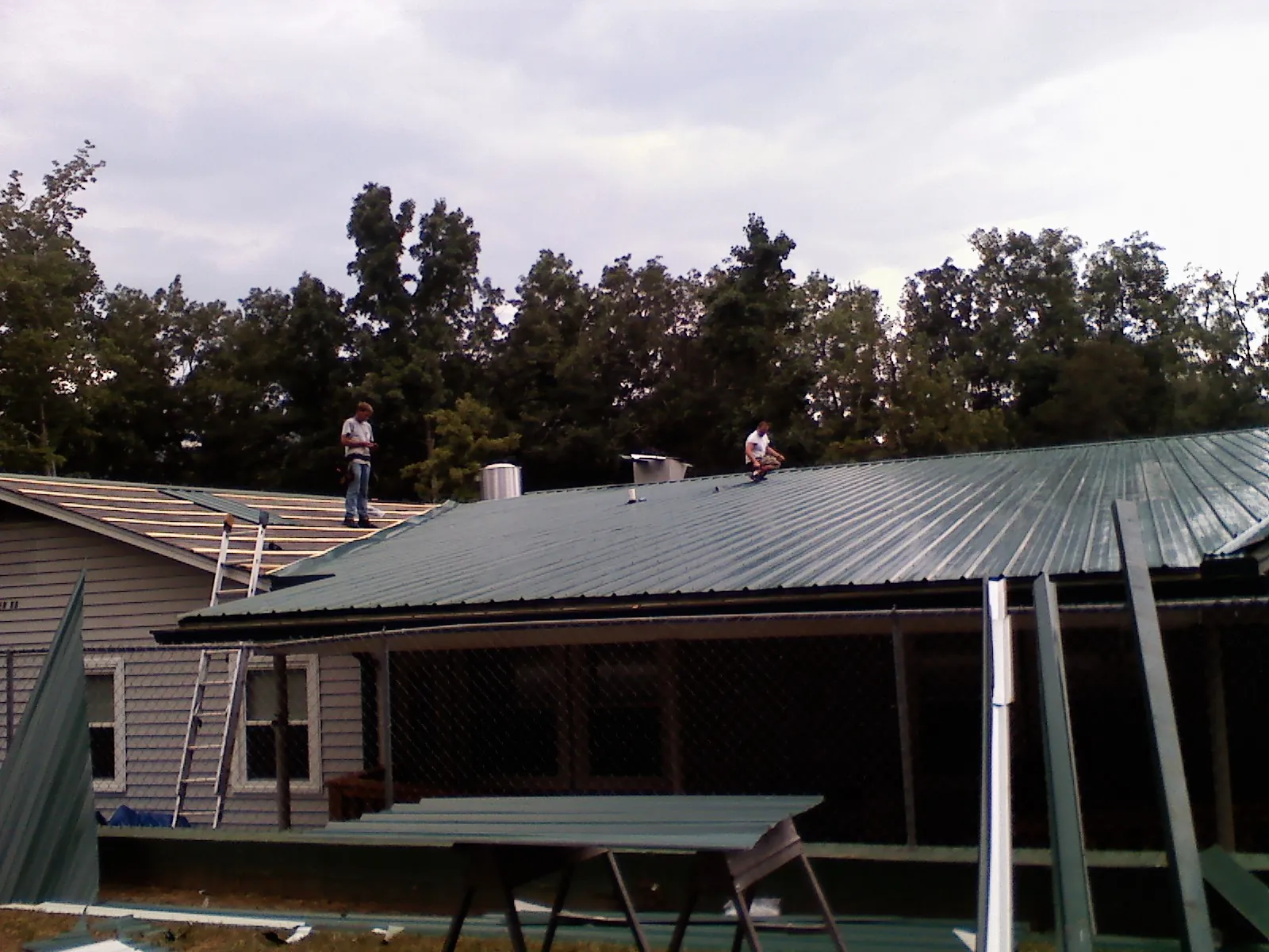 Two roofers installing green metal panels above a covered porch in Snover